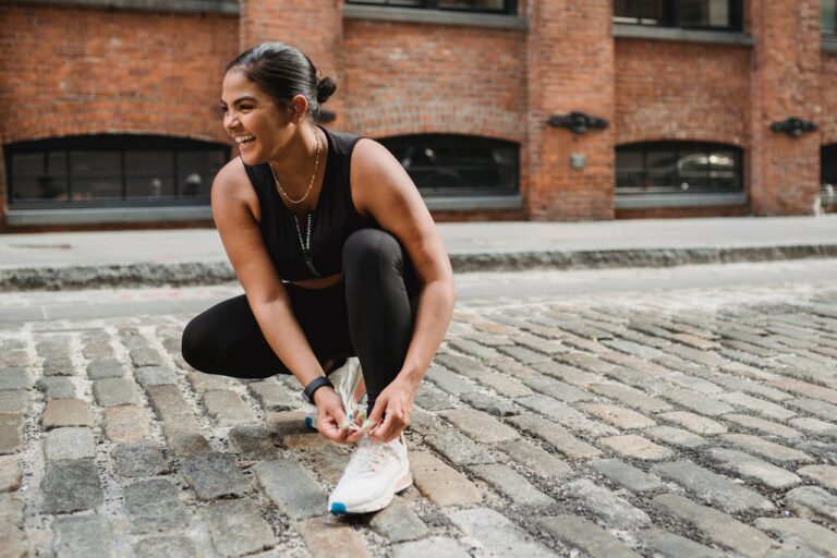 Smiling woman in athletic wear tying sneakers on a cobblestone street outdoors.